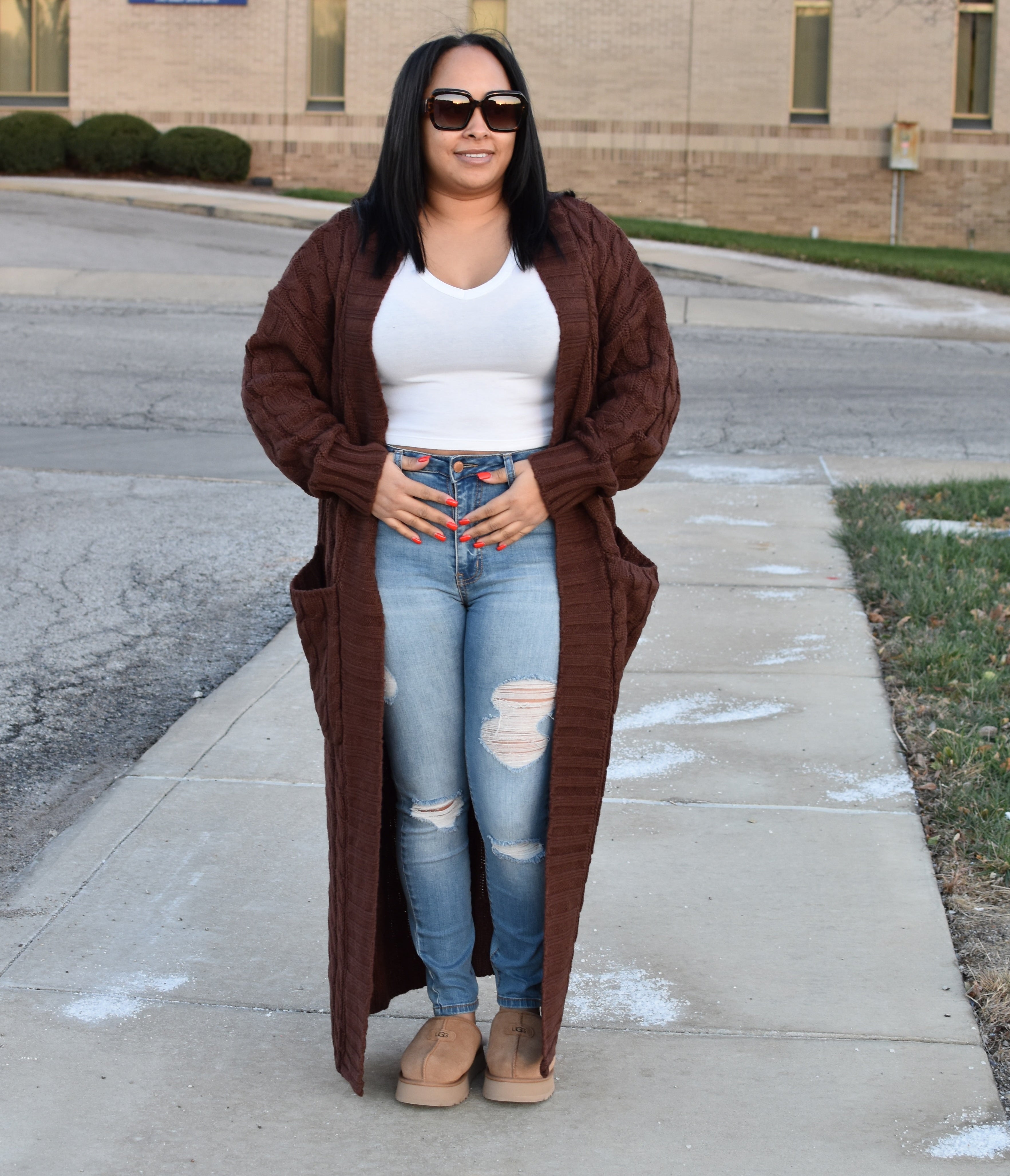Woman wearing a long brown cardigan, white top, and blue jeans standing on a sidewalk.
